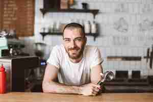 Free photo smiling barista leaning on bar counter and happily looking in camera while holding portafilter in hand in cafe