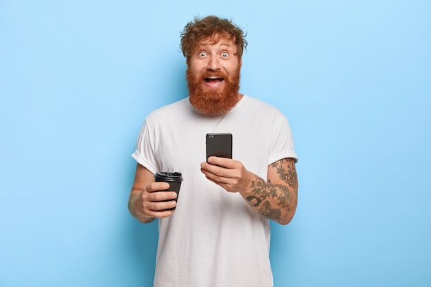 Smiling bearded emotional man has red hair, holds mobile phone, shares great news with friend, gazes with broad smile and bugged eyes, wears casual white t shirt, holds takeaway coffee