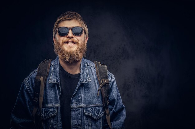Smiling bearded hipster guy wearing sunglasses and backpack dressed in jeans jacket. Isolated on a dark background.