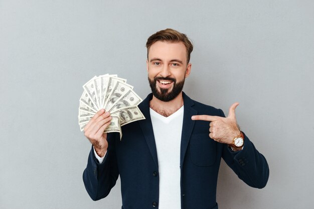 Smiling bearded man in busines clothes holding money and pointing on their while looking at the camera over gray 