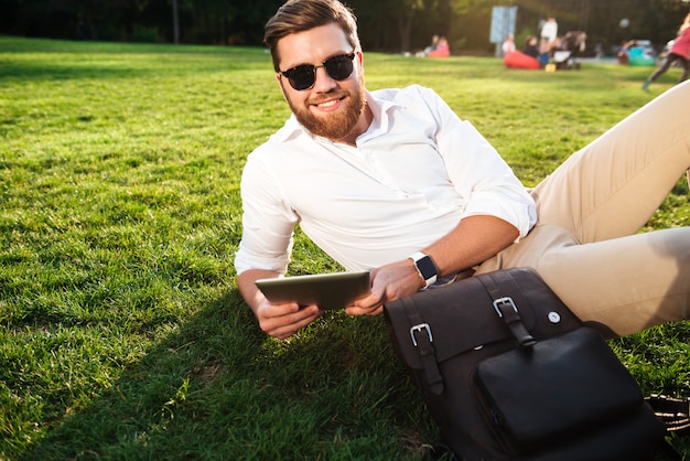 Улыбающийся бородатый мужчина в темных очках, лежа на траве на открытом воздухе с планшетного компьютера и смотрит в камеру