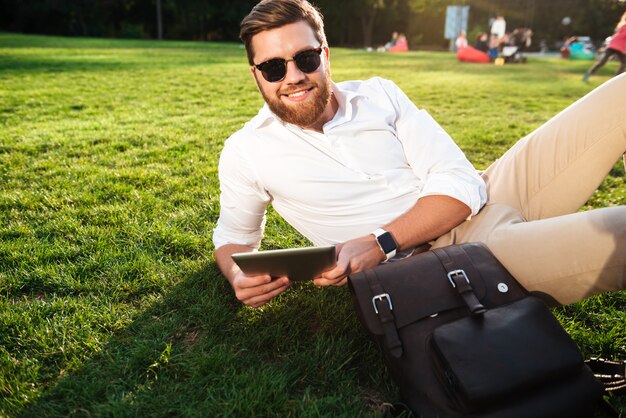 タブレットコンピューターで屋外の芝生の上に横たわって、カメラを見てサングラスのひげを生やした男の笑顔
