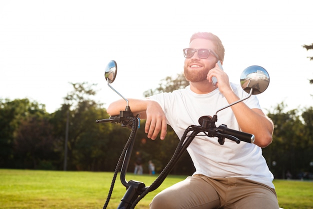 Улыбающийся бородатый мужчина в темных очках сидит на современном мотоцикле на свежем воздухе и разговаривает по смартфону