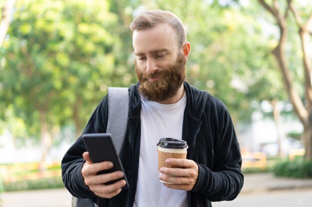 Smiling bearded man walking in city and using smartphone