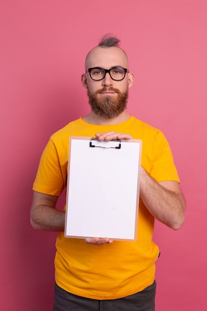 Foto gratuita sorridente uomo barbuto che indossa occhiali da vista che indossano abiti casual in possesso di un blocco per appunti con una carta bianca che punta