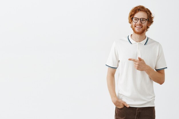 Smiling bearded redhead guy posing against the white wall with glasses