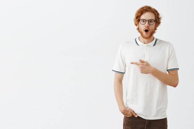 Smiling bearded redhead guy posing against the white wall with glasses