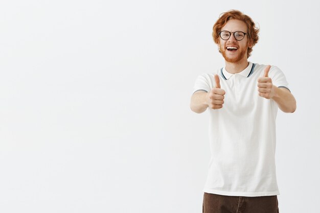 Smiling bearded redhead guy posing against the white wall with glasses