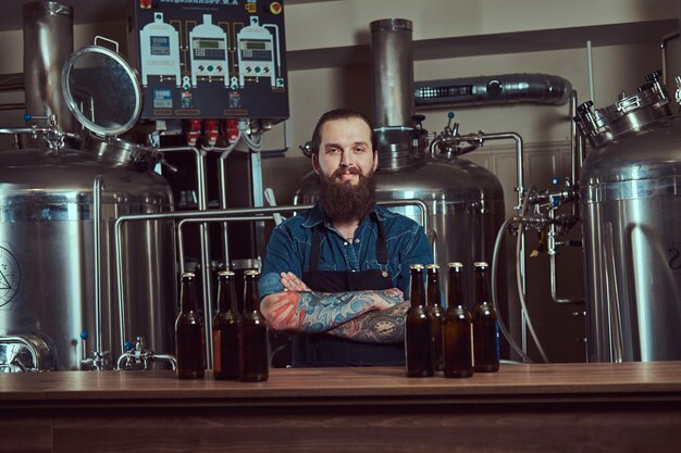 Улыбающийся бородатый татуированный хипстер в джинсовой рубашке и фартуке, работающий на пивоваренном заводе, стоит со скрещенными руками за прилавком.