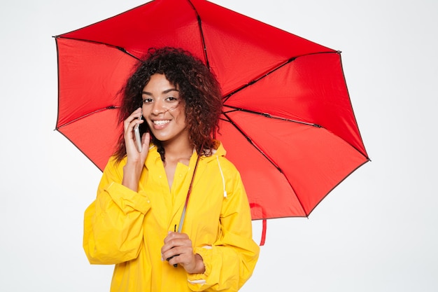 Smiling beautiful african woman in raincoat hiding under umbrella while talking by smartphone and looking at the camera over white background