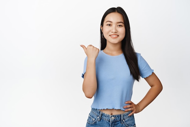 Бесплатное фото Улыбающаяся красивая азиатская девушка, указывающая влево, выглядит уверенно и счастливо, показывая способ продажи баннера, стоящего в футболке на белом фоне