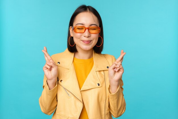 Smiling beautiful asian woman wishing cross fingers for good luck and looking hopeful standing over blue background Copy space