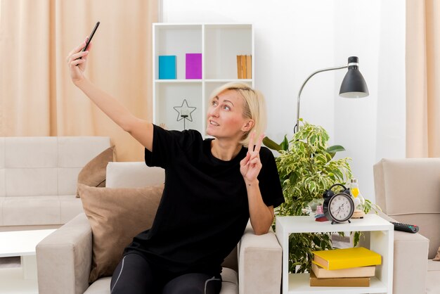 Smiling beautiful blonde russian woman sits on armchair gesturing victory hand sign looking at phone taking selfie inside living room