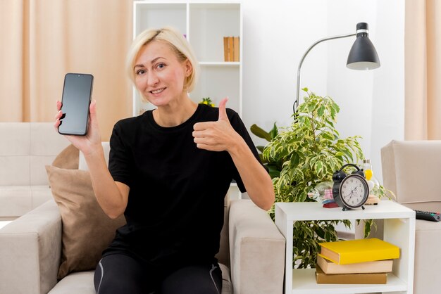 Smiling beautiful blonde russian woman sits on armchair holding phone and thumbs up inside living room