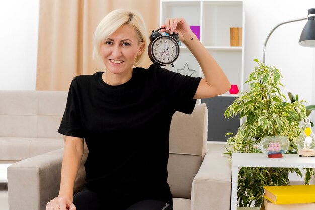 Smiling beautiful blonde woman sits on armchair holding alarm clock