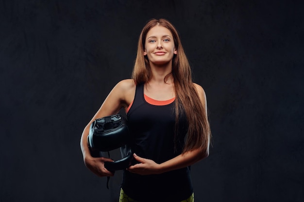 Бесплатное фото Улыбающаяся красивая брюнетка-боксер в спортивной одежде держит защитный шлем и смотрит в камеру. изолированные на темном текстурированном фоне.