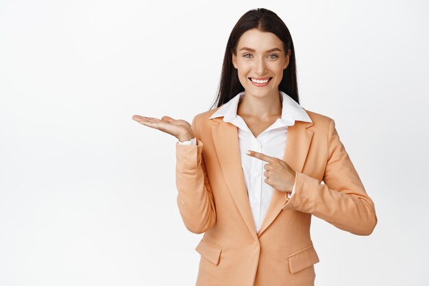 Smiling beautiful corporate woman pointing at her hand holding something showing product on empty palm standing in suit over white background