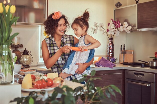 Улыбающаяся красивая кудрявая латиноамериканская мать учит свою милую маленькую дочь готовить пиццу на кухне.