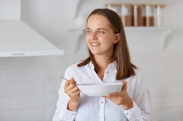 Smiling beautiful dark haired female wearing white shirt, looking smiling away with pleasant smile, posing in light kitchen at home, holding plate in hand.
