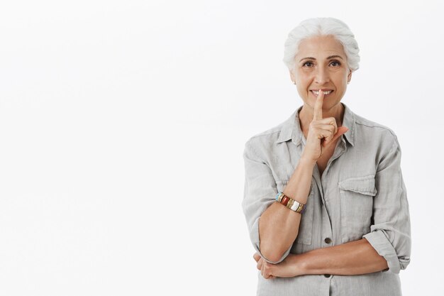 Smiling beautiful elder woman showing shush gesture, tell secret, hushing over white background