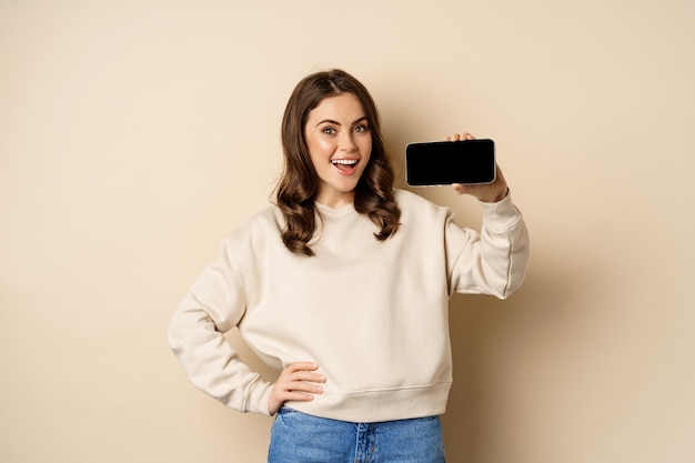 Smiling beautiful female model demonstrating horizontal smartphone screen mobile application standin...