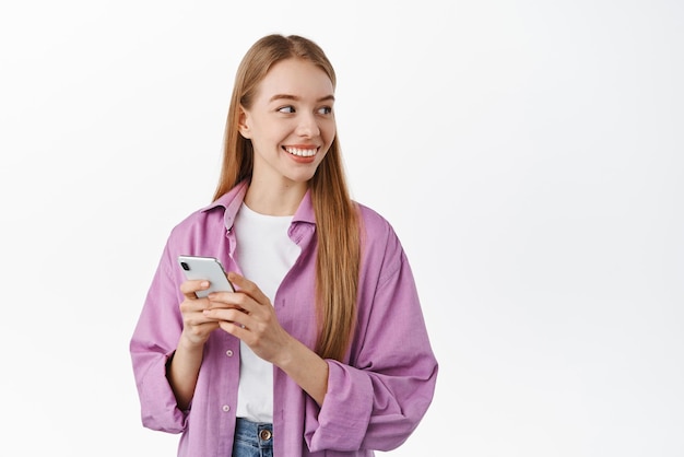 Smiling beautiful female model looking aside at banner logo on copyspace holding smartphone chatting or online shopping standing against white background