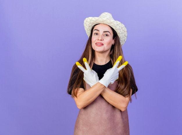 Sorridente bella ragazza giardiniere in uniforme indossando guanti e cappello da giardinaggio attraversando le mani e mostrando il gesto di pace isolato su priorità bassa blu