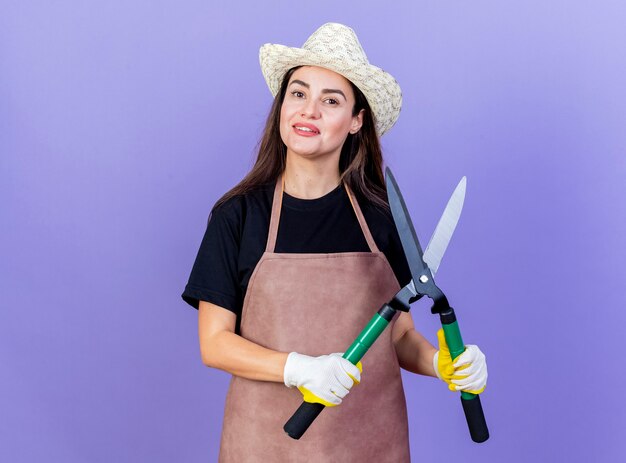 青い背景で隔離のクリッパーズを保持しているガーデニング帽子と手袋を身に着けている制服を着た美しい庭師の女の子の笑顔