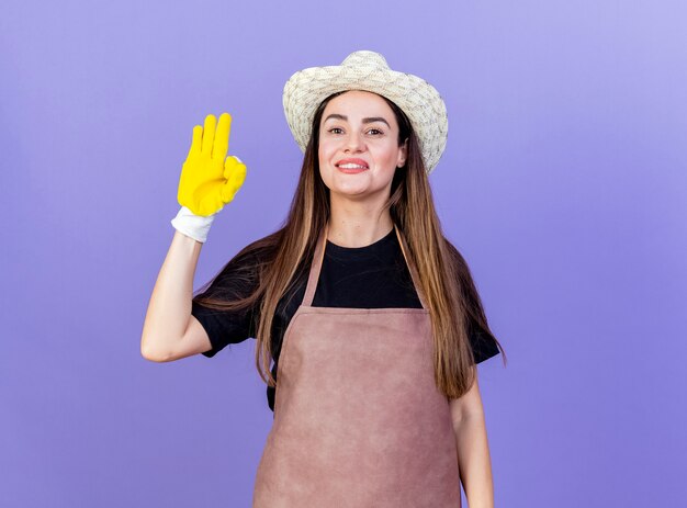 Smiling beautiful gardener girl in uniform wearing gardening hat and gloves showing okey gesture isolated on blue