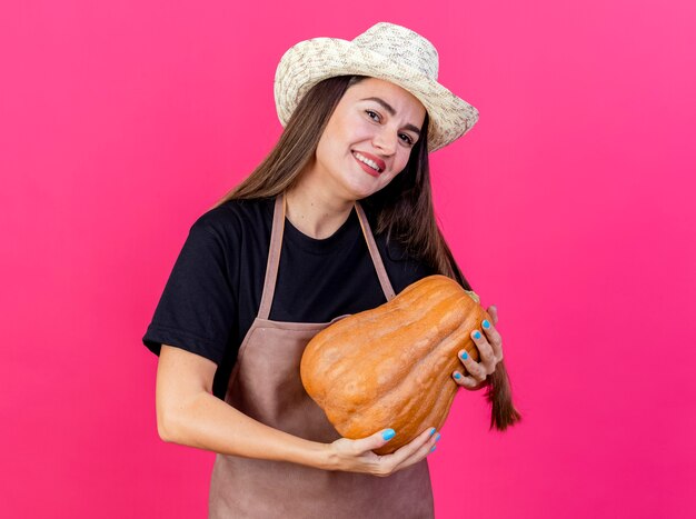 Smiling beautiful gardener girl in uniform wearing gardening hat holding pumpkin isolated on pink background