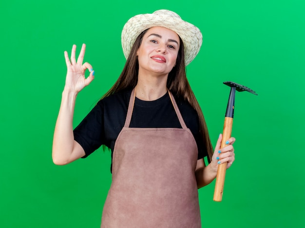 Foto gratuita sorridente bella ragazza giardiniere in uniforme che indossa cappello da giardinaggio tenendo il rastrello e mostrando okey gesto isolato su sfondo verde