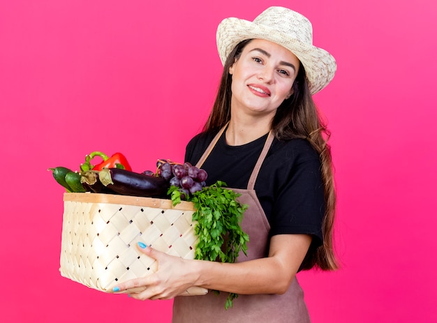 ピンクの背景で隔離の野菜バスケットを保持しているガーデニング帽子を身に着けている制服を着て美しい庭師の女の子の笑顔