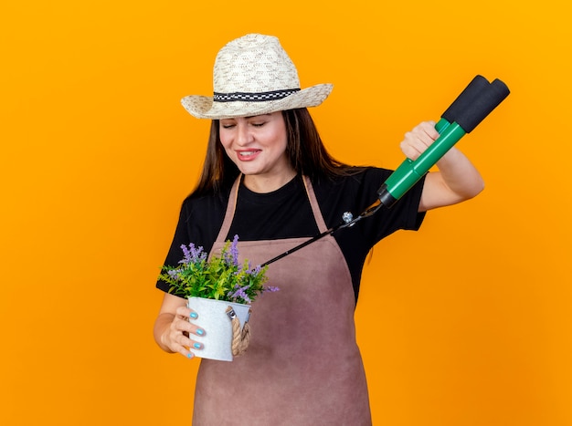 オレンジ色の背景に分離されたクリッパーと植木鉢で花を保持し、切り花の制服と園芸帽子を身に着けている美しい庭師の女の子の笑顔