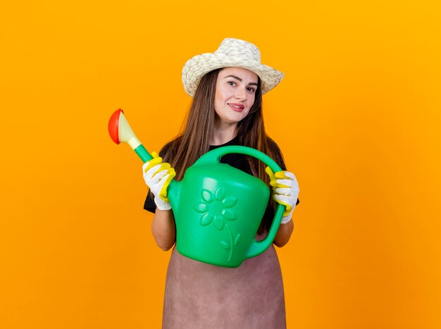 Smiling beautiful gardener girl wearing uniform and gardening hat with gloves holding watering can