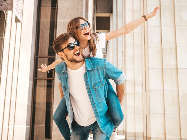 Smiling beautiful girl and her handsome boyfriend in casual summer clothes. Man carrying his girlfriend on the back and she raising her hands. 