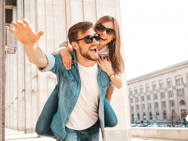 Smiling beautiful girl and her handsome boyfriend in casual summer clothes. Man carrying his girlfriend on the back and she raising her hands.