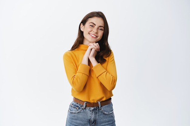 Smiling beautiful girl looking happy and thankful, receiving a gift and admire it, staring at front pleased with hands pressed to chest, white wall