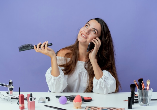 Smiling beautiful girl sits at table with makeup tools holds hair comb talking on phone looking up isolated on purple wall