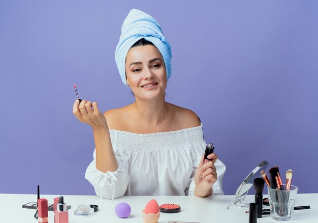 Smiling beautiful girl wrapped hair towel sits at table with makeup tools holding lip gloss looking at side isolated on purple wall