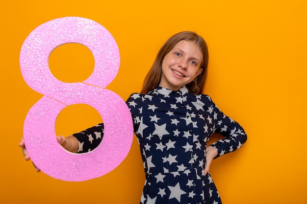 Foto gratuita sorridente bella bambina il giorno della donna felice che porge il numero otto alla telecamera isolata sul muro arancione