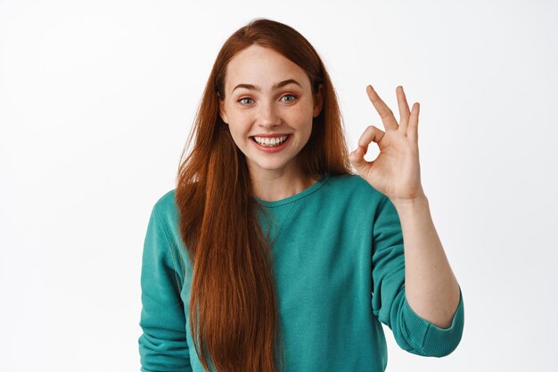 Smiling beautiful redhead girl, shows okay sign, praise and like something good, recommend excellent service, standing satisfied against white background