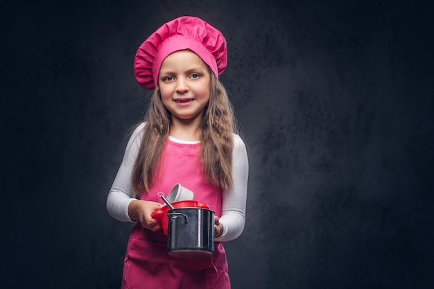 Free photo smiling beautiful schoolgirl dressed in a pink cook uniform holds cookware at a studio. isolated on the dark textured background.