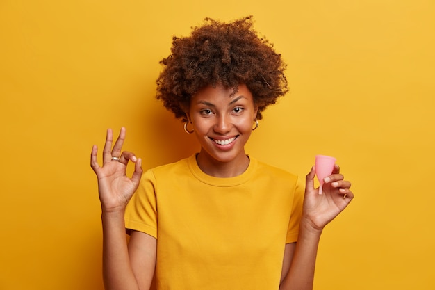 Smiling beautiful woman approves using menstrual cup, makes okay gesture and holds silicone product to insert into vagina in hand gives recommendations for women begginer cup users isolated on yellow