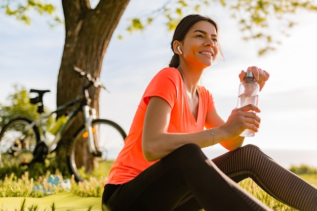 Acqua potabile della bella donna sorridente in bottiglia che fa sport nella mattina nel parco