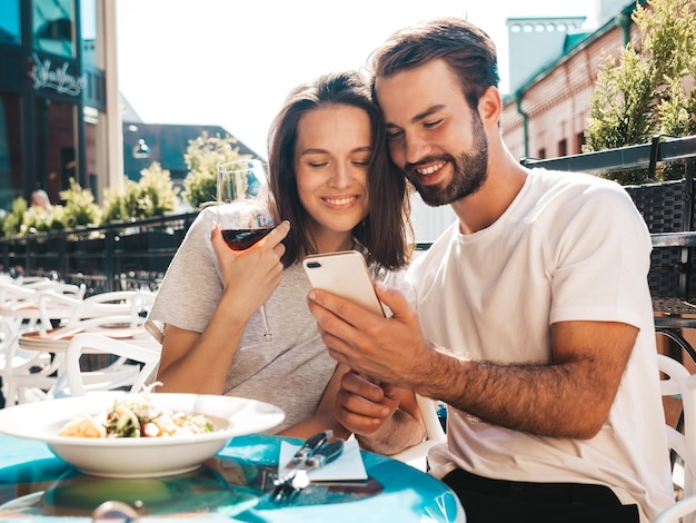 笑顔の美しい女性と彼女のハンサムなボーイフレンドカップルがレストランで彼らのデートで赤ワインのグラスで応援彼らは通りのベランダカフェでアルコールを飲む電話スクリーンを見て