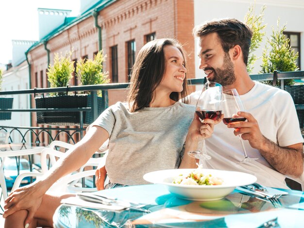 笑顔の美しい女性と彼女のハンサムなボーイフレンド幸せな陽気な家族レストランで彼らのデートで赤ワインのグラスで応援しているカップル彼らは通りのベランダカフェでアルコールを飲んでいます