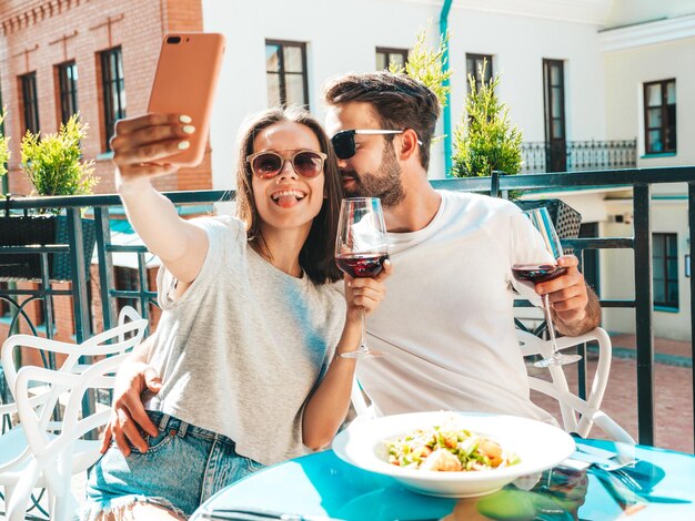 笑顔の美しい女性と彼女のハンサムなボーイフレンド幸せな陽気な家族レストランで彼らのデートで赤ワインのグラスで応援しているカップル彼らは通りのベランダカフェで自分撮りをしています