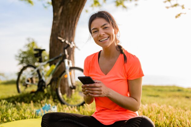Улыбающаяся красивая женщина, держащая телефон, занимается спортом утром в парке