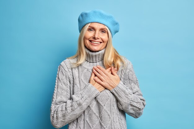 Smiling beautiful woman keeps hands pressed to heart expresses gratitude wears blue beret and knitted grey sweater being thankful and appreciates your help.