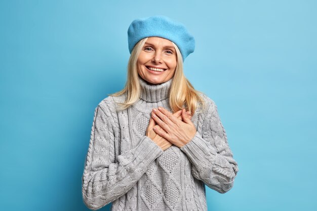笑顔の美しい女性は、手を心に押し付け続け、感謝の気持ちを表し、青いベレー帽と灰色のニットのセーターを着て感謝し、あなたの助けに感謝します。
